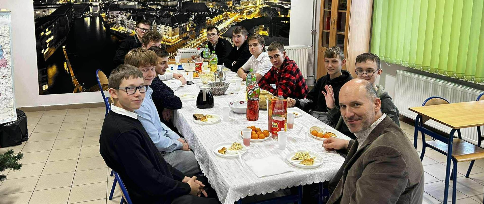 W szkolnej klasie grupa uczniów oraz nauczyciel siedzą przy stole nakrytym białym obrusem, na którym stoją potrawy i napoje.