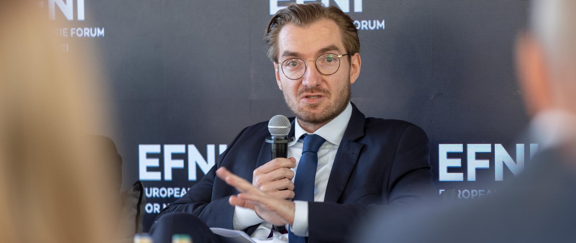 Wiceminister Jan Szyszko siedzi z mikrofonem. Za nim na ściance napis EFNI.