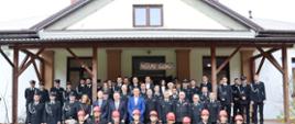 Druhowie OSP strażacy PSP przedstawiciele władzy rządowej i samorządowej oraz młodzież z młodzieżowej drużyny pożarniczej pozują do zdjęcia na tle budynku remizy osp