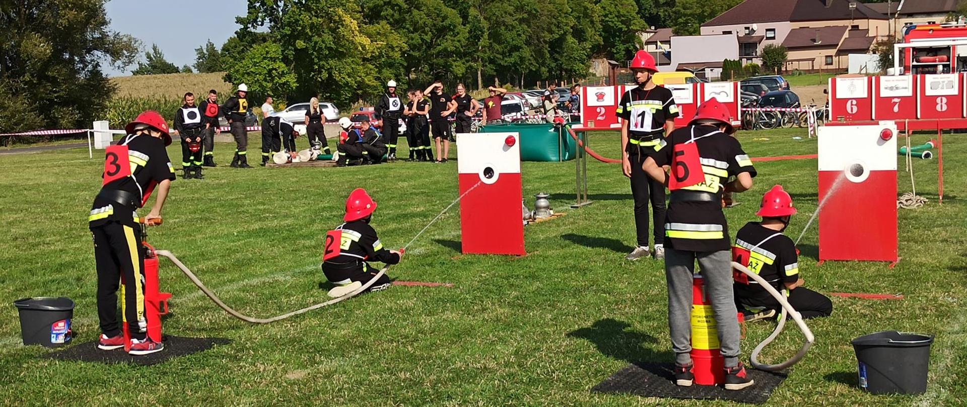 Młodzieżowa drużyna pożarnicza nalewa wodę do biało-czerwonych nalewaków za pomocą hydronetek na płycie boiska. W tle obserwatorzy zawodów, budynek i drzewa.