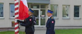 Na zdjęciu widoczny poczet flagowy podczas podnoszenia flagi państwowej na tle budynku Komendy Miejskiej Państwowej Straży Pożarnej. Poczet flagowy składa się z trzech funkcjonariuszy w mundurach galowych.
