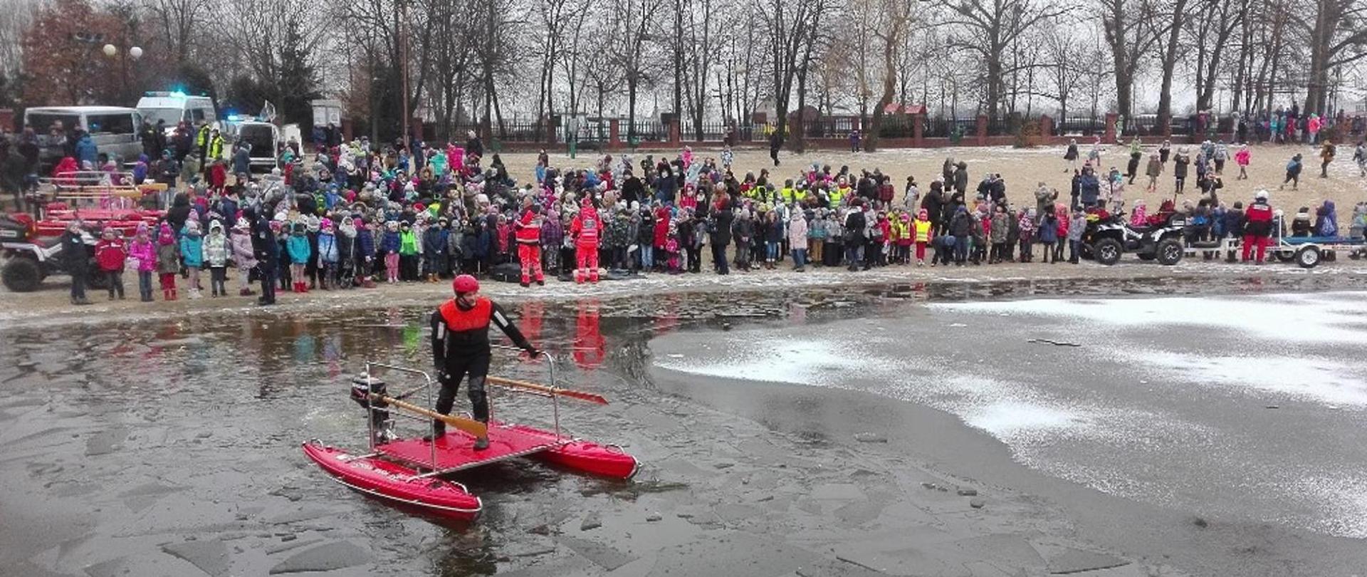 Na pierwszym planie fotografii strażak ubrany w kombinezon wypornościowy, porusza się przy pomocy deski wodno lodowej po pokruszonym lodzie. Na drugim tle osoby postronne oglądający pokaz działań - ćwiczeń wodno lodowych.