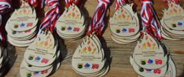 Zawody sportowo-pożarnicze. Na stoliku eksponowane są okolicznościowe medale dla zawodników z MDP. Maja kształt płomyka. Widnieje na nich napis oraz herb Gminy Rawicz, logo ZOSP RP i MDP. 