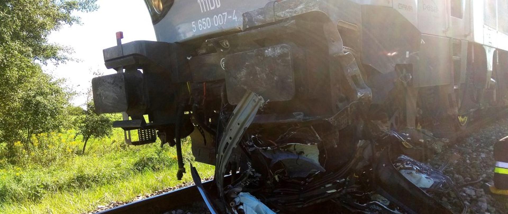 Zdjęcie przedstawia przód niebiesko – popielatej lokomotywy pociągu pośpiesznego stojącej na torach. Pod lewym jej zderzakiem, w kierunku pierwszego lewego koła, znajdują się duże fragmenty pogniecionej karoserii samochodu, której część spoczywa na kamiennym nasypie.Za lokomotywą, na drugim lanie widoczne są fragmenty łąki i rosnących drzew.
