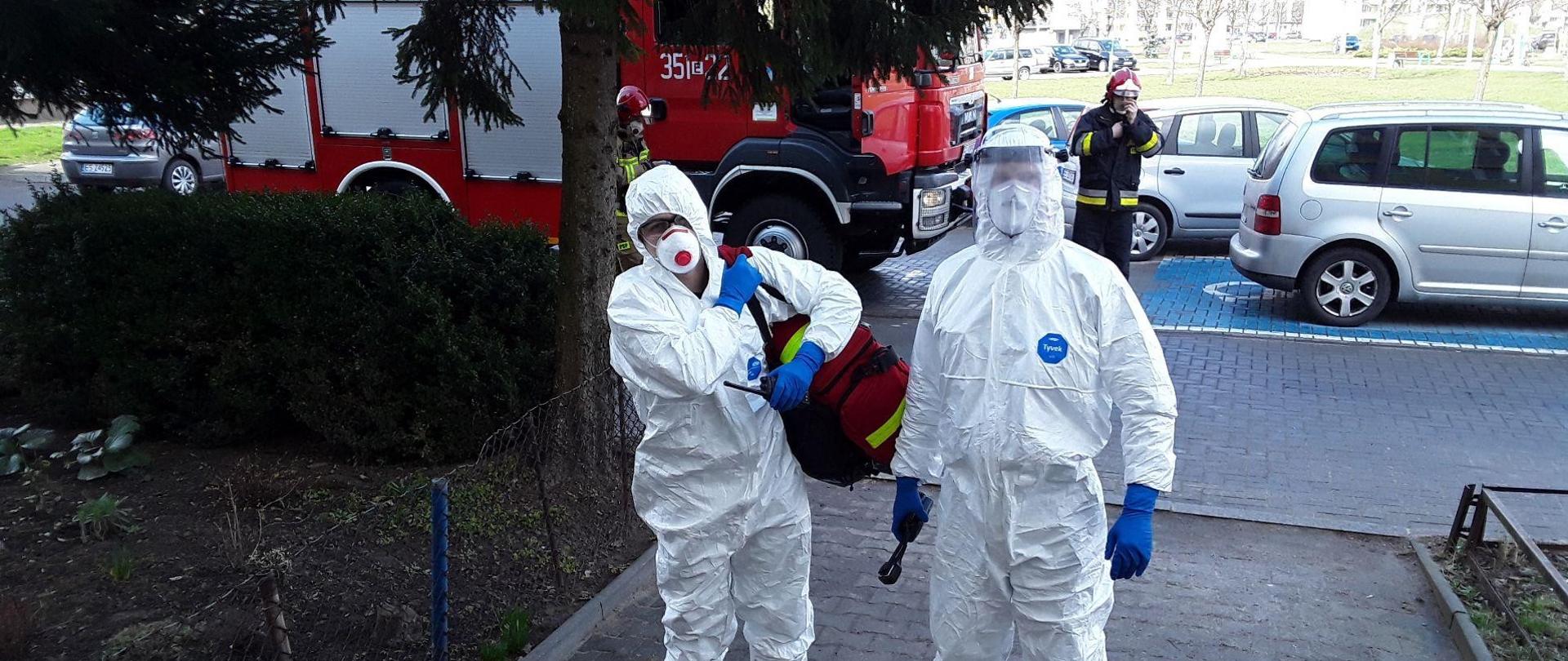 Fotografia przedstawia strażaków państwowej straży pożarnej przygotowanych do udzielenia pierwszej pomocy osobie z koronawirusem ubranych w specjalne ubrania ochrony biologicznej ze sprzętem medycznym na tle pojazdu pożarniczego i innych strażaków. 