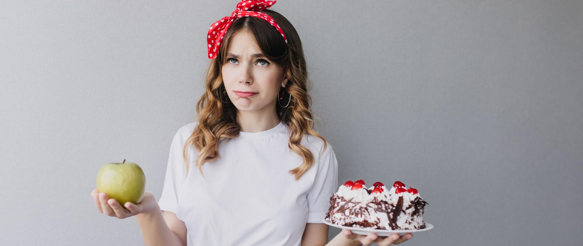 Dziewczyna blondynka w kręconych włosach, białym T-shircie i biało-czerwonej spódnicy stoi i myśli o swojej diecie. W swojej prawej dłoni trzyma zielone jabłko, a w lewej dłoni trzyma talerzyk ze śmietanowym tortem i wiśniami.
