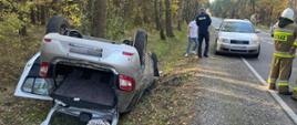 Na zdjęciu widoczny samochód osobowy leży na dachu na poboczu drogi, częściowo zanurzony w rowie, z otwartym bagażnikiem i drzwiami. Obok stoją funkcjonariusze policji i strażak, a także inne osoby. Na drodze, po prawej stronie, zaparkowany jest srebrny samochód. W tle widoczny jest las.