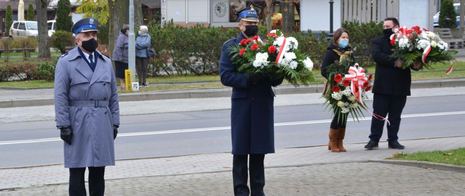 Zdjęcie przedstawia komendanta powiatowego Policji oraz komendanta powiatowego Państwowej Straży Pożarnej w Ustrzykach Dolnych trzymającego kwiaty. Za nimi na prawo widać kobietę i mężczyznę którzy również trzymają kwiaty. Za nimi jest droga i chodnik na którym stoją dwie kobiety i rozmawiają. W oddali widać budynki 