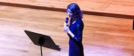 Kobieta stojąc przy pulpicie na estradzie sali koncertowej mówi do mikrofonu.