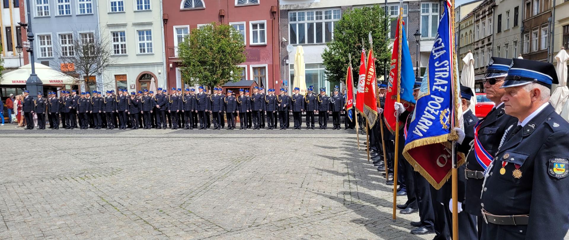 Zdjęcie przedstawia pododdziały strażackie stojące podczas uroczystego apelu na wybrukowanym placu w dwuszeregu podczas zbiórki. Po lewej strażacy stoją w dwuszeregu. Po prawej w szeregu ze sztandarami. W tle fronty zabytkowych kamienic.