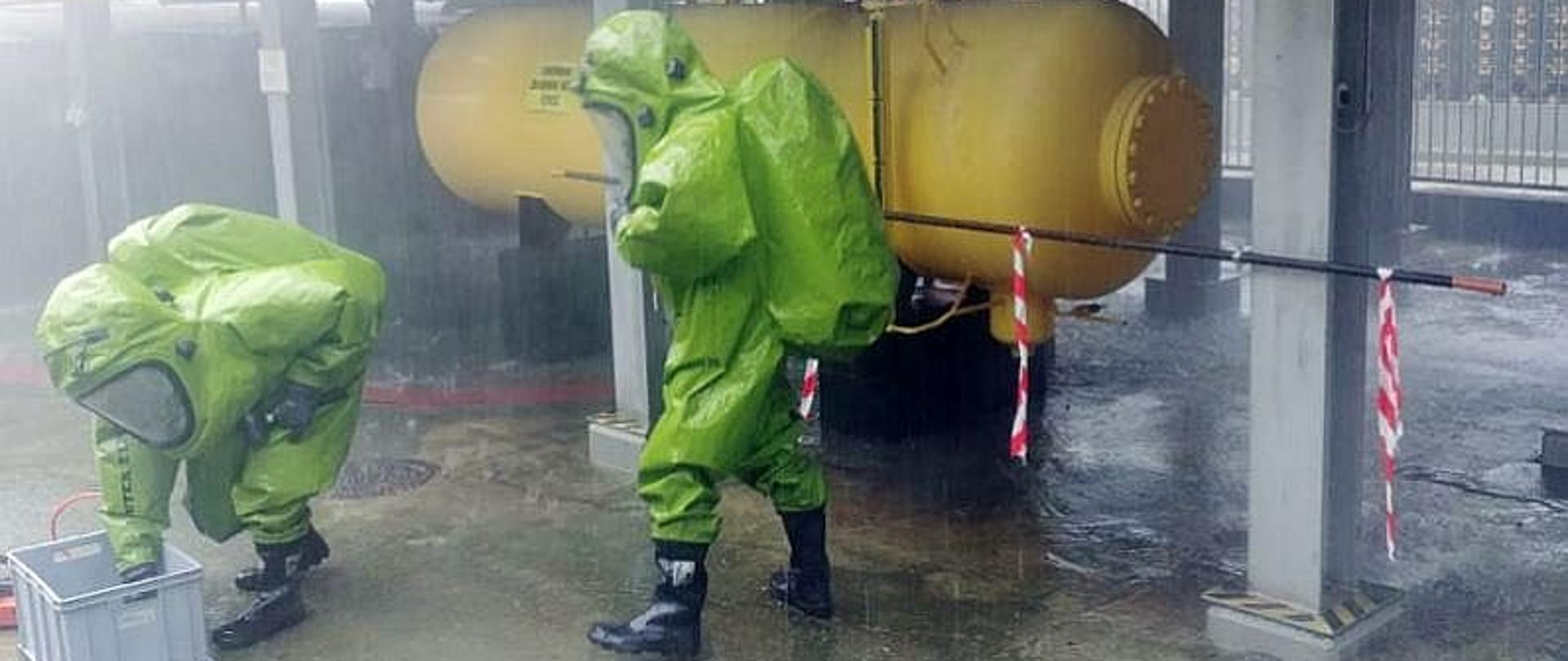 Dwóch ratowników podczas ćwiczeń z uszczelniania instalacji z amoniakiem. Strażacy ubrani są w chemoodporne ubrania chemiczne koloru zielonego, stoją na tle żółtego zbiornika z amoniakiem.