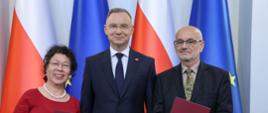 Prezydent Duda stoi pomiędzy mężczyzną w czarnym garniturze i kobietą w czerwonej sukience, za nimi flagi Polski i UE.