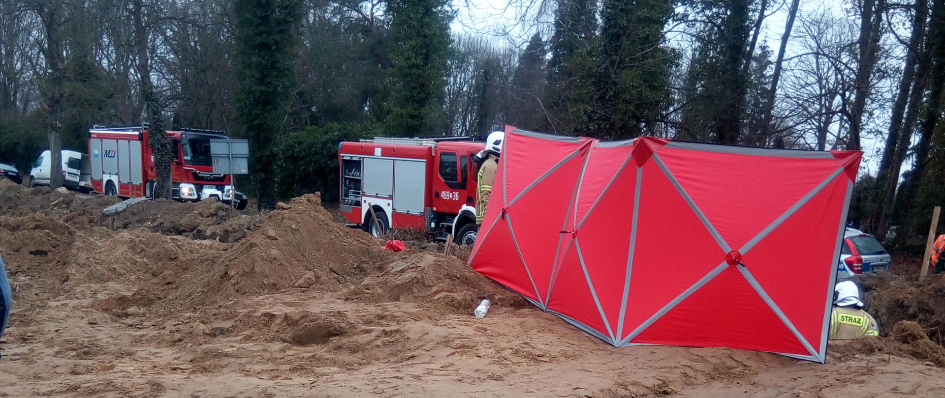 Zdjęcie przedstawia miejsce wypadku na budowie gdzie całe zdarzenie zasłonięte jest parawanem Straży Pożarnej a w tle na drodze stoją pojazdy ciężarowe PSP