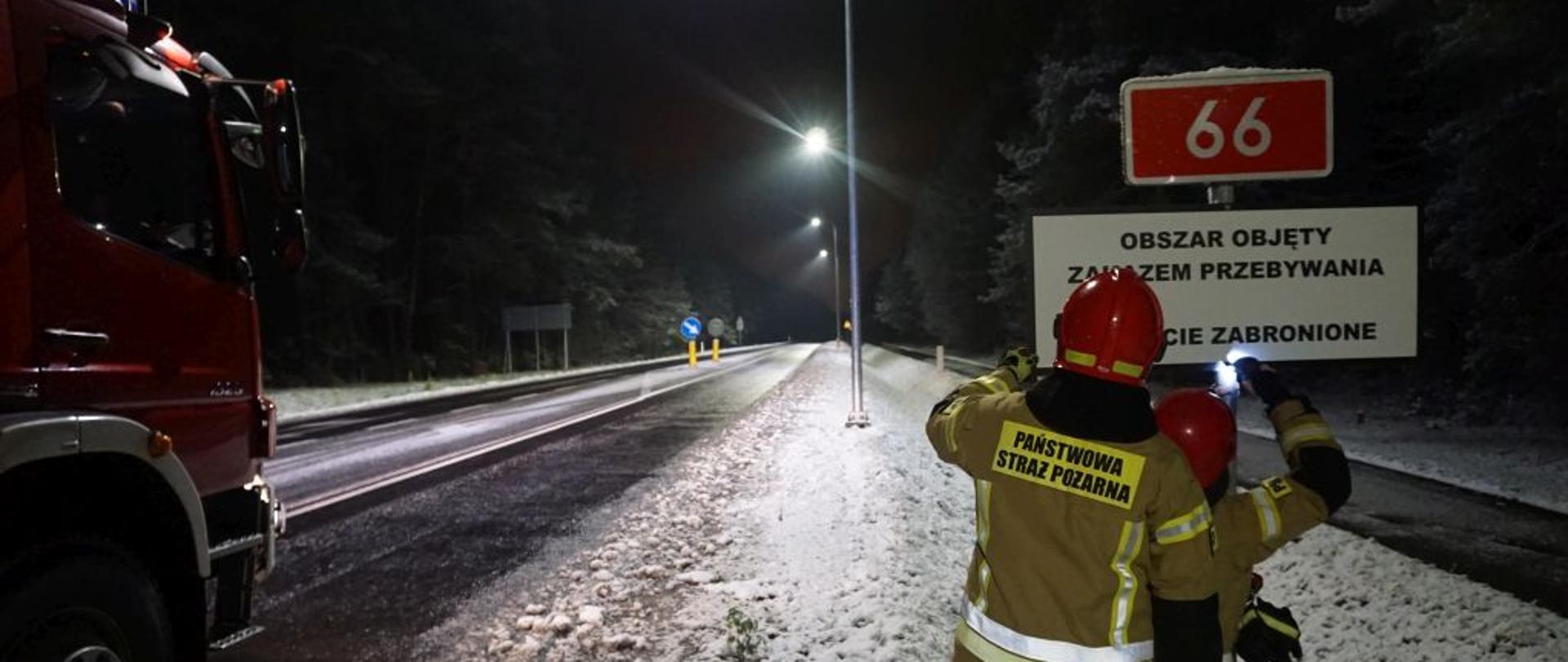 Na zaśnieżonej drodze strażacy obok czerwonego wozu bojowego przykręcają tablicę informacyjną.