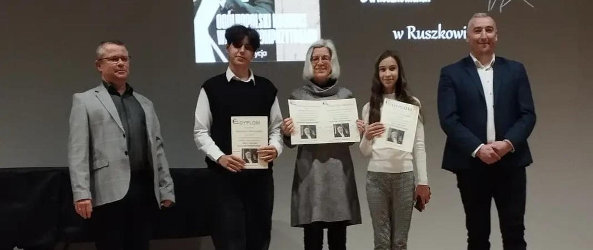 Na zdjęciu widać uczniów którzy trzymają dyplomy w rękach. P koło nich stoją nauczyciele a w tle widoczne jest wyświetlany slajd na którym jest nazwa szkoły muzycznej oraz zdjęcie Pendereckiego.