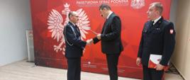 Zdjęcie przedstawia ministra, który wręcza decyzję o przyznaniu świadczenia ratowniczego druhowi OSP. Na zdjęciu widać również komendanta. Osoby stoją przed banerem komendy.