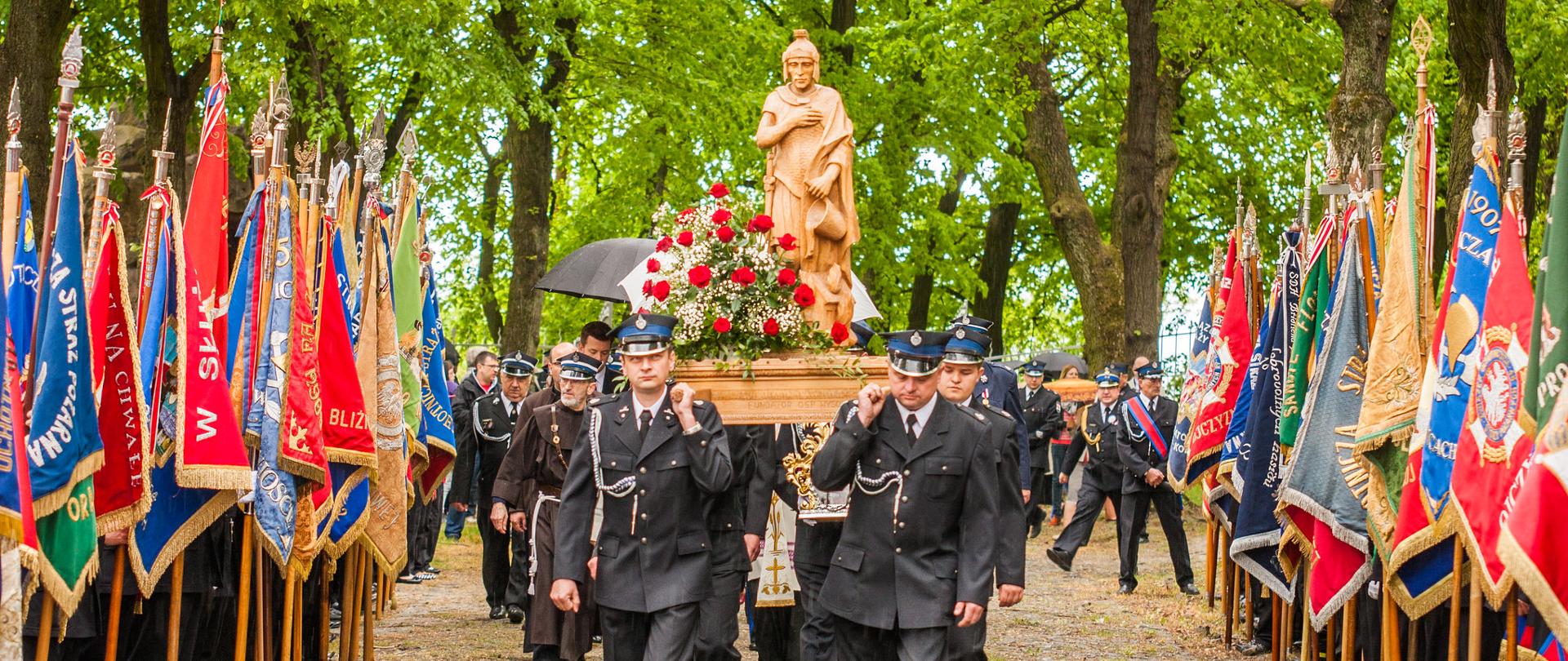Uroczystości w ramach pielgrzymki strażaków do Sanktuarium Świętej Anny Samotrzeciej na Górze Świętej Anny. Strażacy ochotnicy w mundurach galowych niosą na barkach drewnianą figurę Świętego Floriana. Po lewej i prawej stronie widoczne poczty sztandarowe OSP. W tle widoczne drzewa.