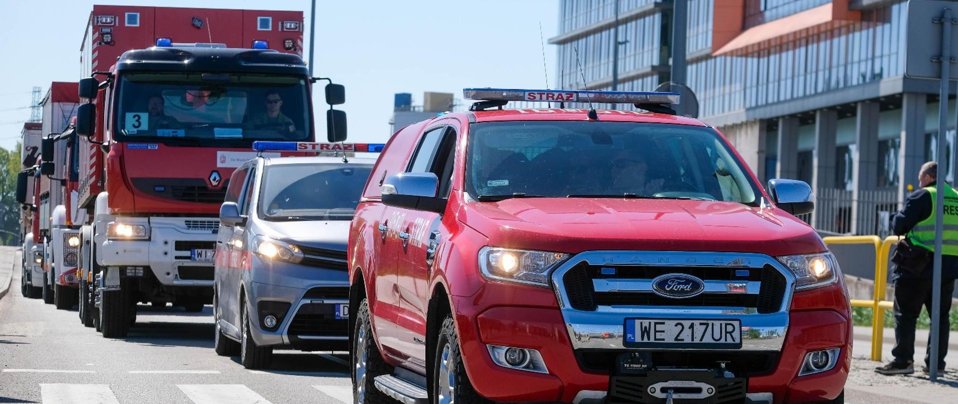 Kolumna pojazdów Państwowej Straży Pożarnej przemieszczająca się przez miasto; na czele czerwony Ford Ranger z sygnalizacją świetlną, za nim pojazdy operacyjne i ciężarowe z modułami kontenerowymi. W tle nowoczesne zabudowania miejskie.