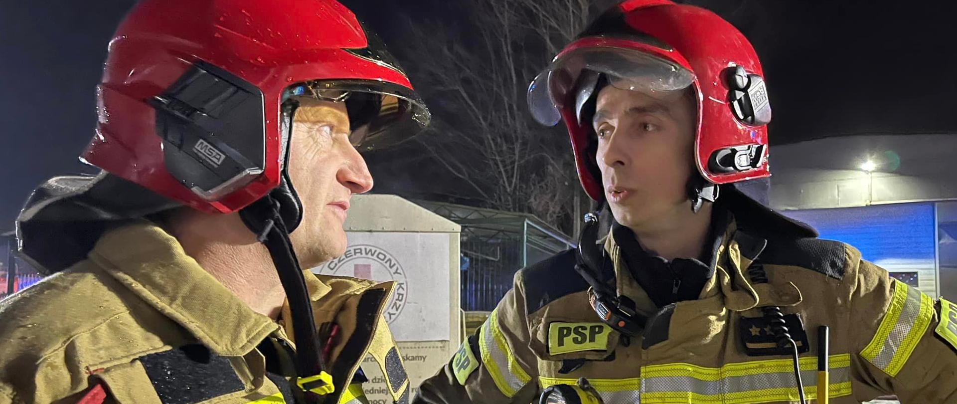 Dwóch strażaków w mundurach specjalnych, z hełmami na głowach - dyskutują o działaniach ratowiczo-gaśniczych. W tle obiekty garażowe, płot, drzewa. Pora po zmierzchu.