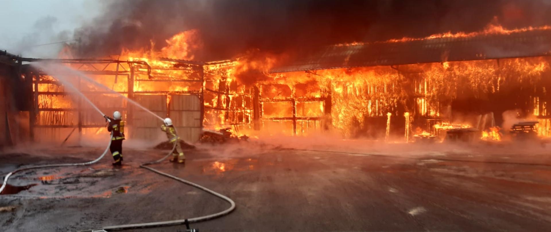 Zdjęcie przedstawia pożar tartaku. Jeden z budynków całkowicie objęty pożarem. Na zdjęciu widać również strażaków, którzy podają wodę na palący się budynek. 
