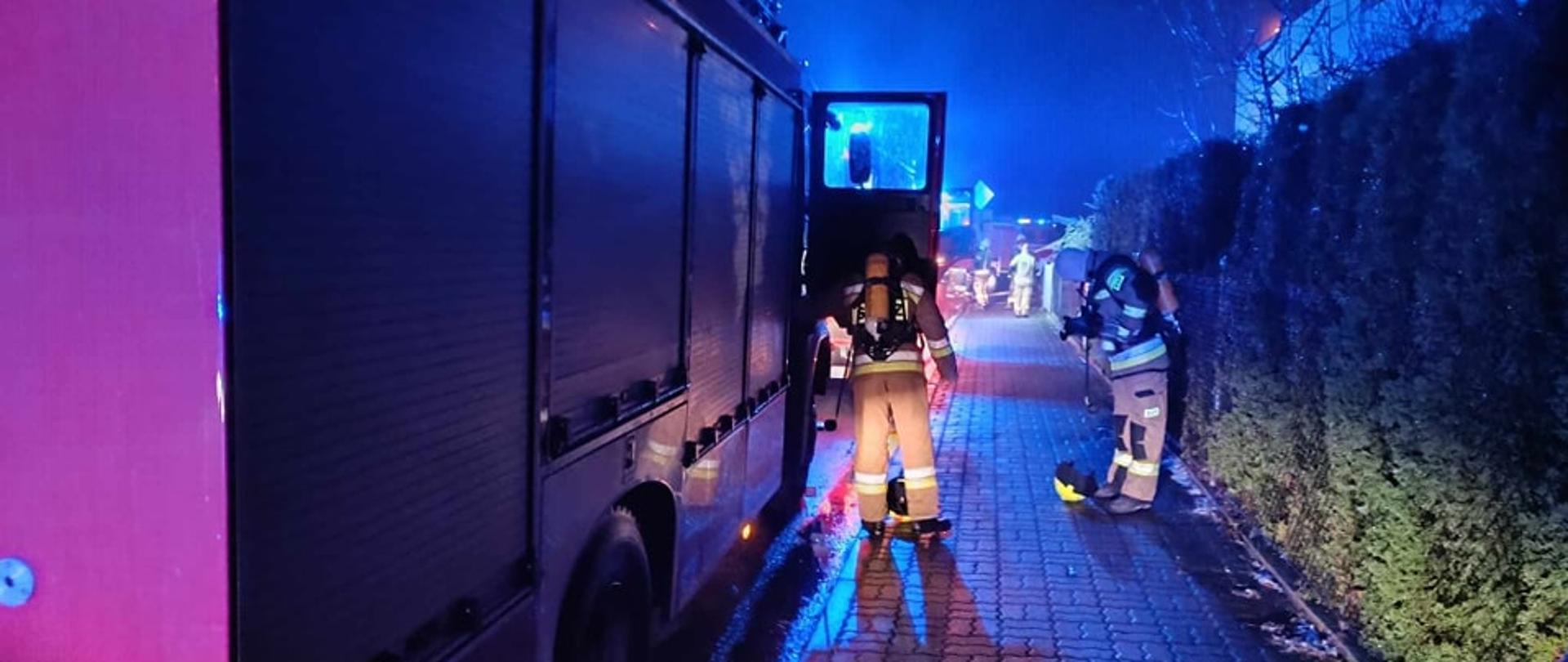 Samochody strażackie z włączonymi sygnałami świetlnymi. Obok strażacy zakładają aparaty powietrzne. 