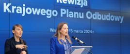 Minister funduszy i polityki regionalnej Katarzyna Pełczyńska-Nałęcz stoi przy pulpicie i przemawia do mikrofonu, obok niej stoi tłumaczka języka migowego, za ich plecami grafika na niebieskim tle z informacją o rewizji KPO
