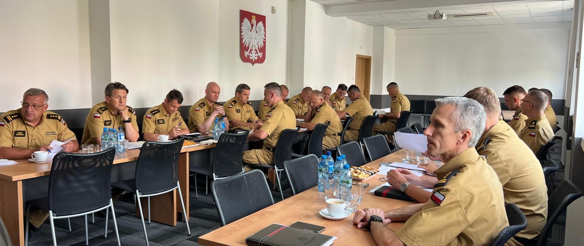 Na zdjęciu uczestnicy narady siedzą w sali. Wszyscy to strażacy w piaskowych mundurach. Na ścianie sali zawieszone godło RP (Biały orzeł w złotej koronie)