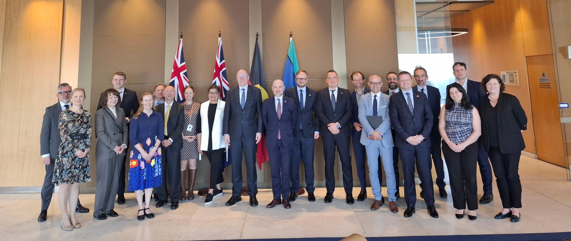 Uczestnicy polskiej delegacji stoją na tle flag. 