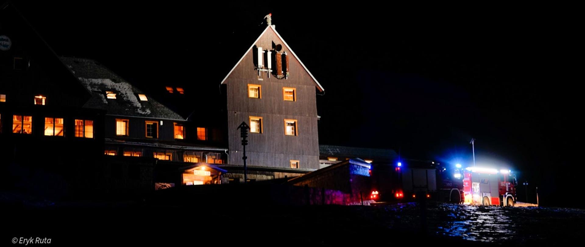 Schronisko na Hali Szrenickiej podczas działań gaśniczych w dniu 11 listopada 2021 roku. W tle budynek schroniska o zmroku a przed nim dwa pojazdy straży pożarnej z włączoną sygnalizacją alarmową.