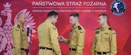 Czterech mężczyzn w beżowych mundurach strażackich stoi przed czerwonym tłem z napisem „Państwowa Straż Pożarna – Komenda Wojewódzka PSP w Warszawie”. Jeden z mężczyzn wręcza drugiemu małą złotą statuetkę. Pozostali obserwują ceremonię. Na tle widoczne jest też godło Polski i logo mazowieckiej komendy PSP.