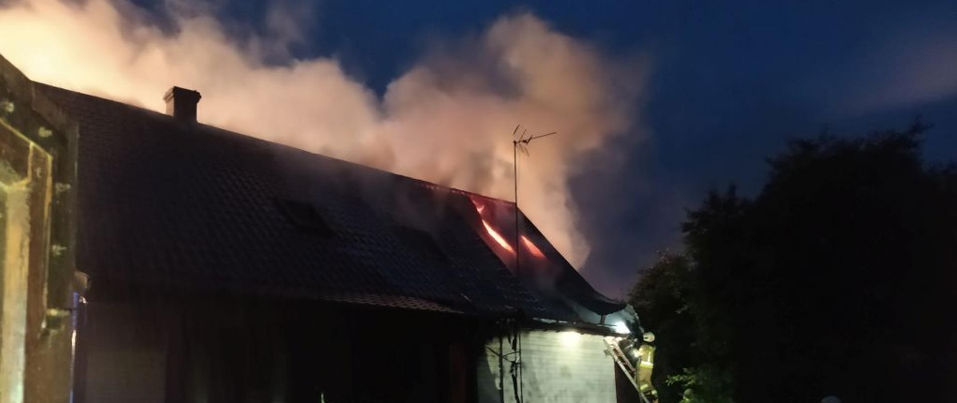 W nocy. Budynek jednorodzinny biały. Przez blaszane pokrycie wydobywają się płomienie. Jeden ze strażaków stoi na drabinie opartej o dach budynku. Trzech strażaków w ciemnych mundurach bojowych prowadzi akcję gaśniczą.