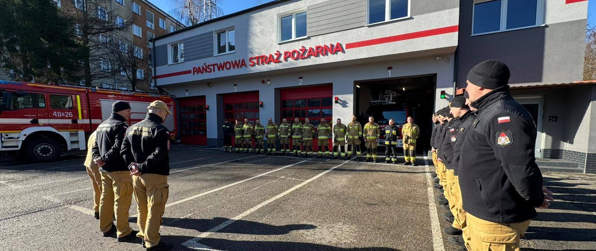 Plac wewnętrzny Jednostki ratowniczo Gaśniczej. Na placu grupa strażaków z prawej w czarnych polarach a na wprost w mundurach bojowych. W tle budynek komendy i garaże bojowe.