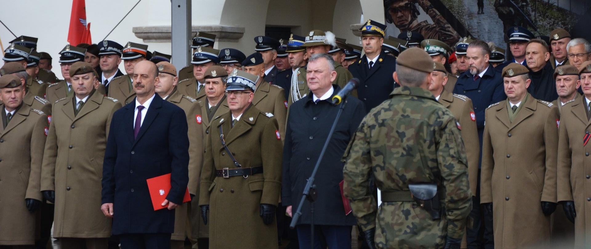 Trybuna honorowa podczas apelu WOT na Rynku Wielkim w Zamościu; szeregi oficerów w umundurowaniu galowym, w tle baner z żołnierzami.”