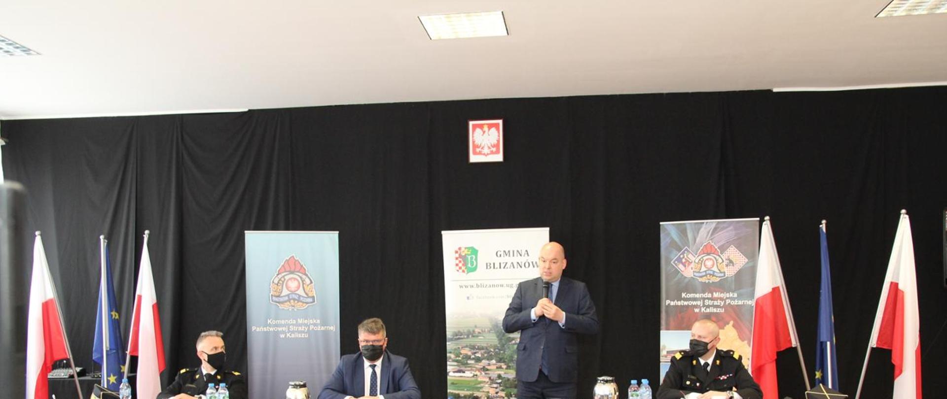 Na zdjęciu widzimy czterech mężczyzn siedzących przy stole. Pierwszy z lewej to strażak ubrany w galowy mundur koloru ciemnego, na ramionach widzimy żółte dystynkcje, na twarzy maseczka koloru czarnego. Kolejny mężczyzna od lewej to cywil ubrany w szary garnitur, białą koszulę, ciemny krawat, czarna maseczka na twarzy, okulary na oczach. Kolejny Mężczyzna to również cywil, który stoi przy stole i w rękach trzyma mikrofon. Ubrany jest w szary garnitur, niebieską koszulę. Ostatni mężczyzna to strażak ubrany w galowy mundur koloru ciemnego, na ramionach widzimy żółte dystynkcje, na twarzy maseczka koloru czarnego. Za mężczyznami widzimy flagi Polski oraz Unii Europejskiej. Dalej widzimy ciemne tło, na którym powieszone jest godło Polski oraz trzy banery przedstawiające Komendę miejską Państwowej Straży Pożarnej w Kaliszu oraz Gminę Blizanów. 