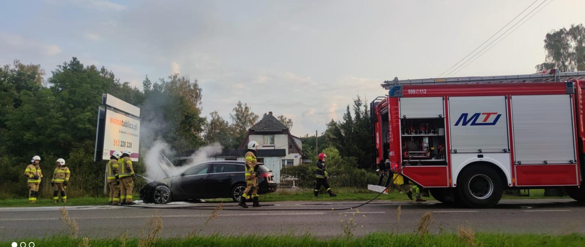 Strażak podaje prąd piany gaśniczej do komory silnika czarnego samochodu. po prawej samochód pożarniczy.