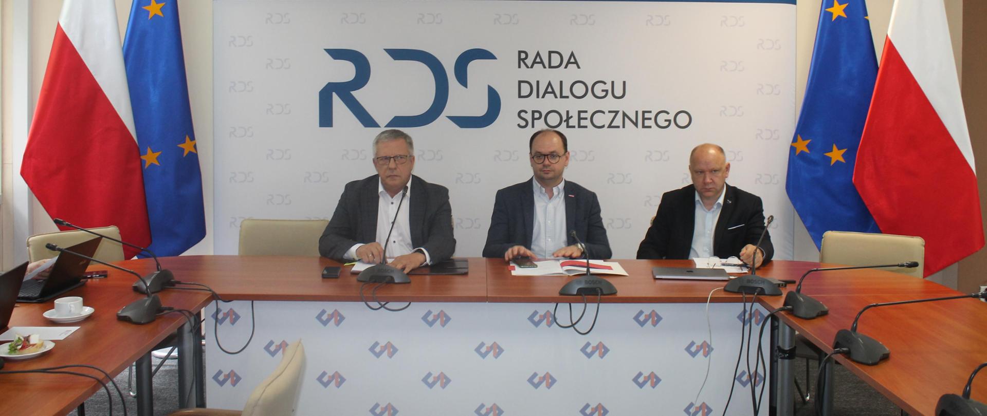Na zdjęciu widnieje stół prezydialny za którym siedzą od lewej strony: Przewodniczący zespołu ds. budżetu, wynagrodzeń i świadczeń socjalnych – Leszek Miętek, Sebastian Gajewski - Podsekretarz Stanu w Ministerstwie Rodziny, Pracy i Polityki Społecznej oraz przedstawiciel Zespołu ds. ubezpieczeń społecznych. Po obu stronach stołu widnieją dwie flagi: Polski i UE. W tle siedzących znajduje się baner z napisem Rada Dialogu Społecznego a nad nim wisi godło Polski.