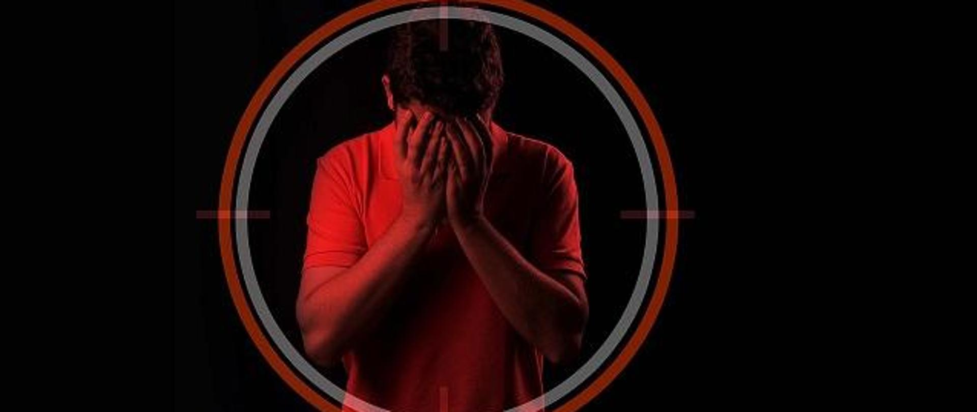 Mężczyzna zasłania twarz rękoma, stoi na czarnym tle, otoczony jest biało-czerwonym okręgiem, przypominającym celownik