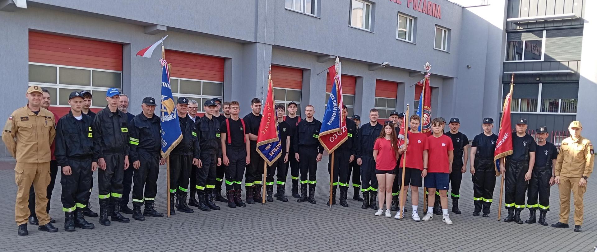 Dwóch wykładowców i grupa strażaków OSP stoi ze sztandarami przed budynkiem KM PSP Konin.