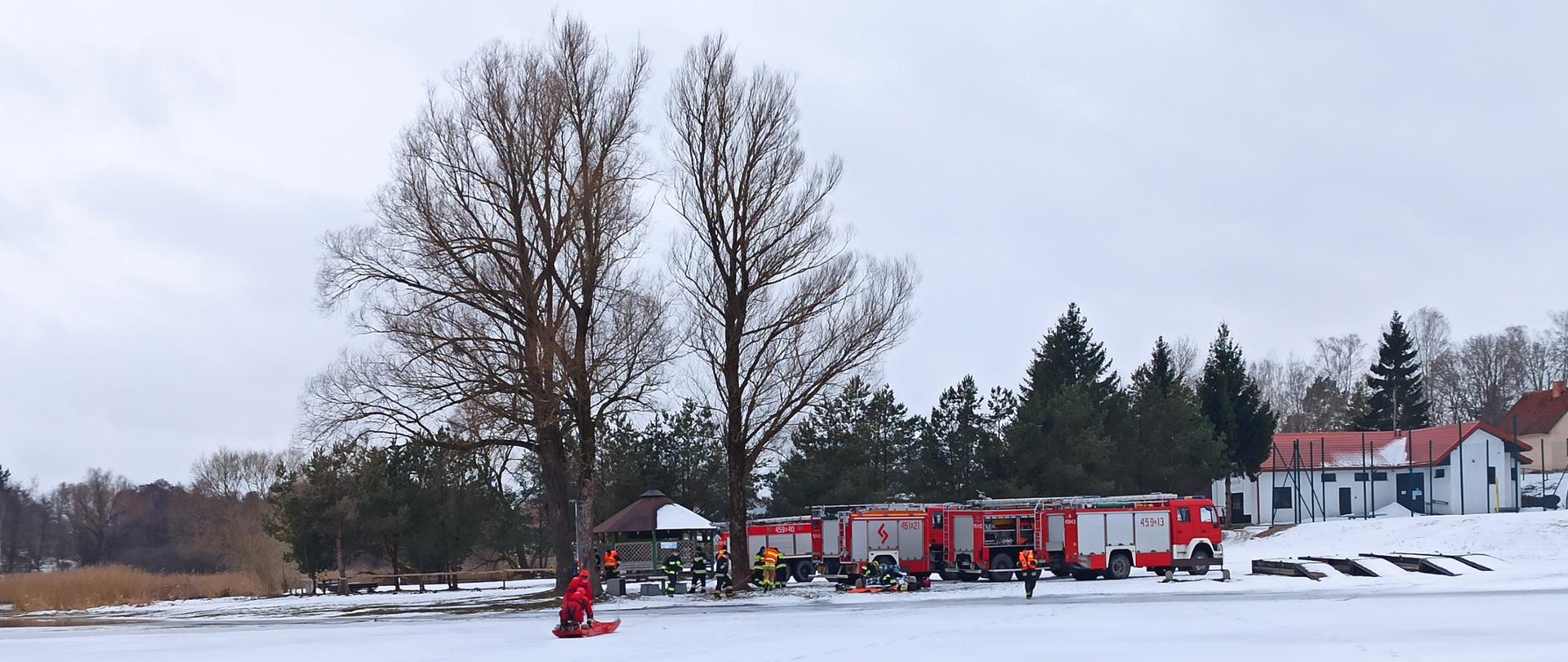 Zdjęcie przedstawia uchwycony z oddali obraz dwóch ratowników na lodzie (odpowiednio zabezpieczonych i wyposażonych w sanie lodowe) na tle strażaków i czterech pojazdów pożarniczych