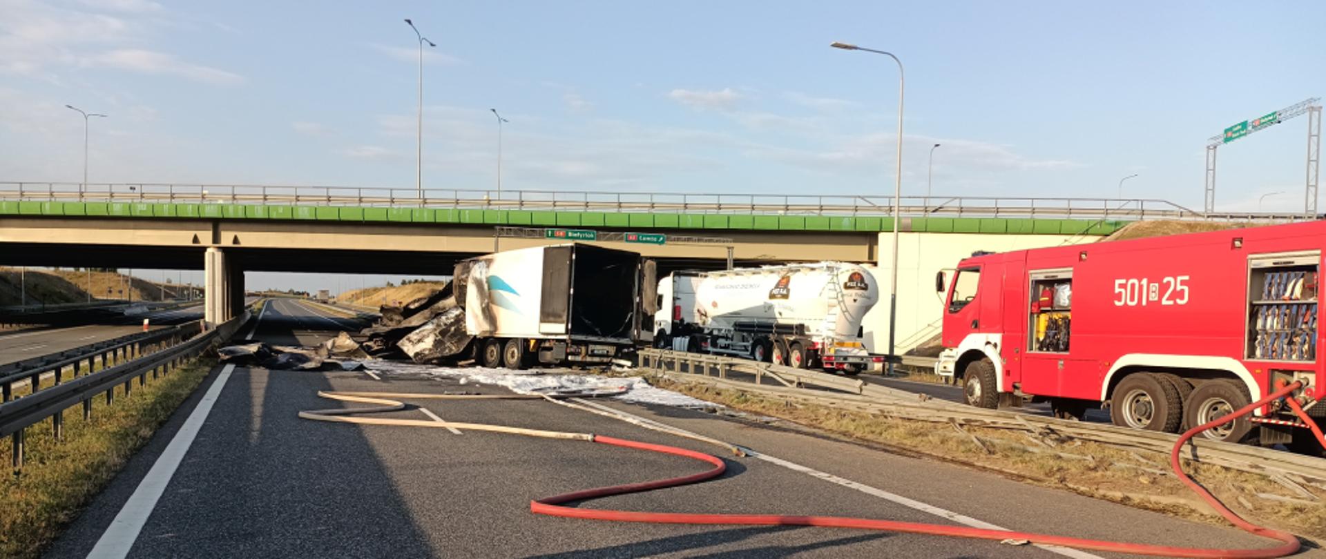 Samochód ciężarowy z całkowicie zniszczonym przodem stojący na kołach na jednym pasie ruchu drogi S8. Samochód nosi znamiona pożaru, przy samochodzie leży wąż strażacki. Za barierkami na drodze stoi wóz strażacki oraz samochód cysterna. Zdjęcie wykonano w słoneczny dzień.