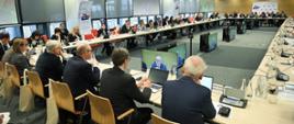 W sali konferencyjnej przy dużym stole siedzi w dwóch rzędach kilkadziesiąt osób i słucha wystąpienia jednego z uczestników spotkania 