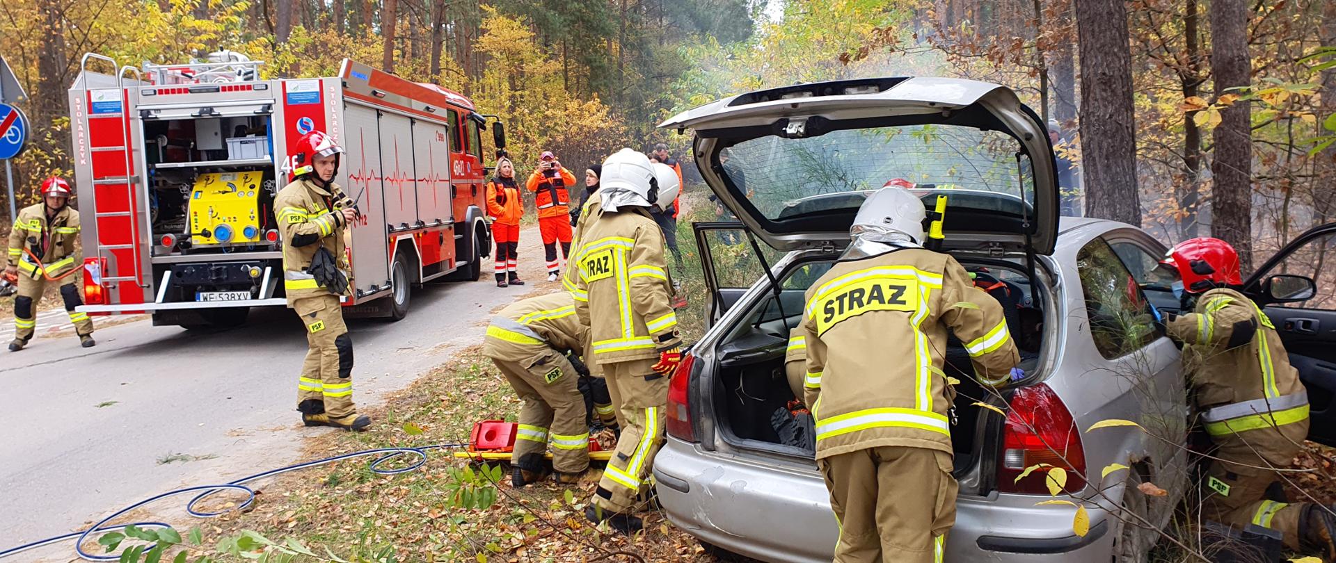 strażacy podczas ćwiczeń na terenie drogi w lesie, czterech strażaków ewakuują poszkodowanego z samochodu po wypadku, dwóch strażaków i dwie osoby ze służb przedmedycznych stoi przy samochodzie pożarniczym