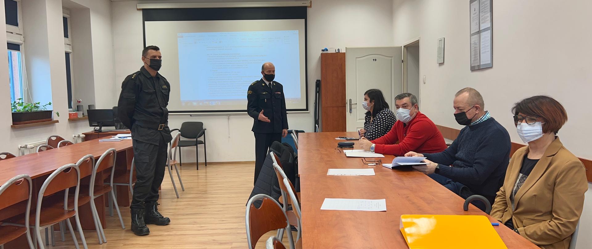 świetlica szkoleniowa, w tle wyświetlane slajdy, po prawej stronie przy stołach siedzące osoby, po lewej stronie dwóch stojących umundurowanych strażaków