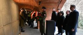 Członkowie Rady Ministrów składają wieniec przed tablicą pamięci. Kwiaty trzyma żołnierz Wojska Polskiego. 