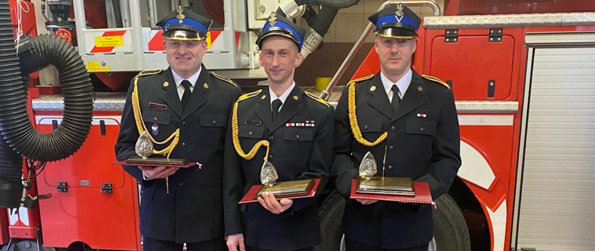 Przejście na zaopatrzenie emerytalne strażaków z podziału bojowego . Strażacy ubrania w ubrania wyjściowe , mundur koloru granatowego ze sznurem i czapką rogatywką. Stoją na tle samochodu specjalnego SHD 23, od lewej strony aspirant Łukasz Lachowski, starszy ogniomistrz Łukasz Dymus i starszy ogniomistrz Sławomir Kowalski
