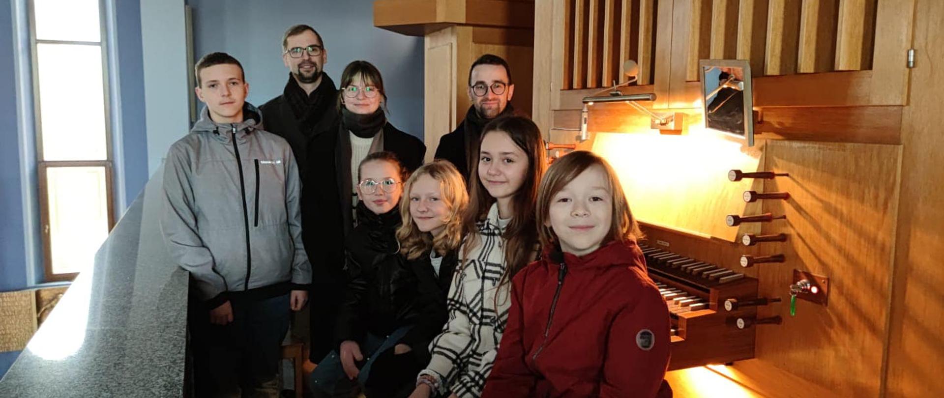 Grupa uczniów i nauczyciele siedzą przed kontuarem organów