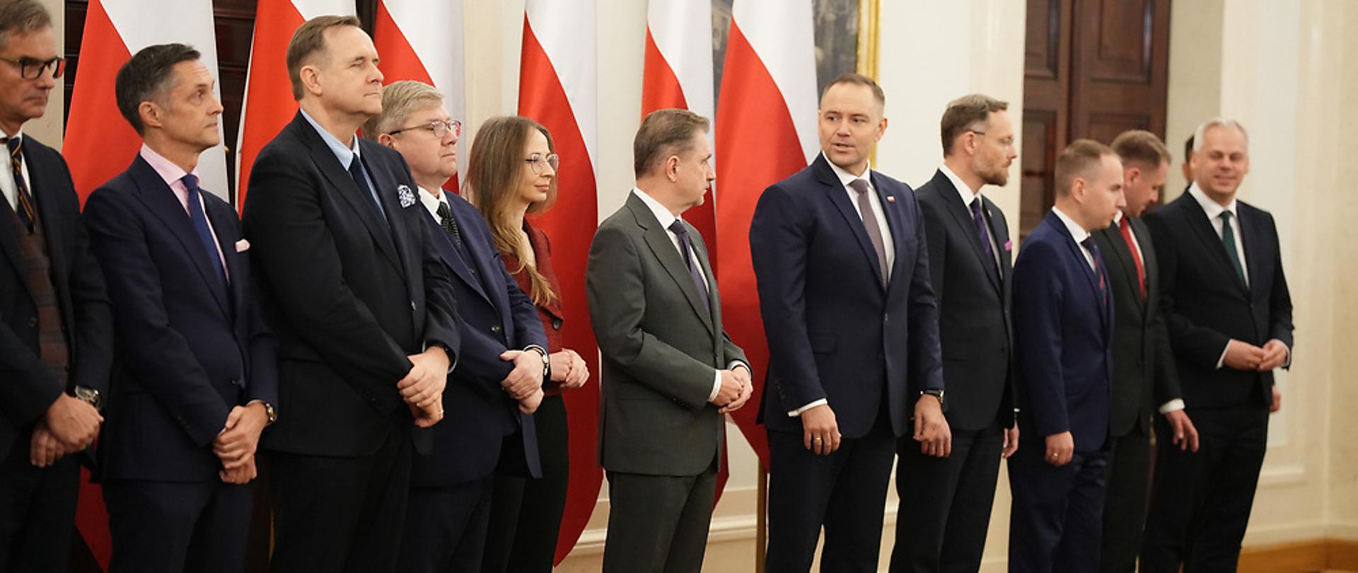 Zdjęcie zrobione w Kancelarii Prezydenta RP gdzie dokonano powołań i odwołań w składzie Rady Dialogu Społecznego. Centralnie stoi Karol Nawrocki – Prezydent RP wraz z przedstawicielami Kancelarii Prezydenta RP po Jego lewej stronie. Po prawej stoją: Piotr Duda – Przewodniczący Rady Dialogu Społecznego, Agnieszka Dziemianowicz-Bąk – Ministra Rodziny, Pracy i Polityki Społecznej, Wiceprzewodnicząca Rady oraz pozostali członkowie Prezydium. W tle stojący pięć flag Polski.
