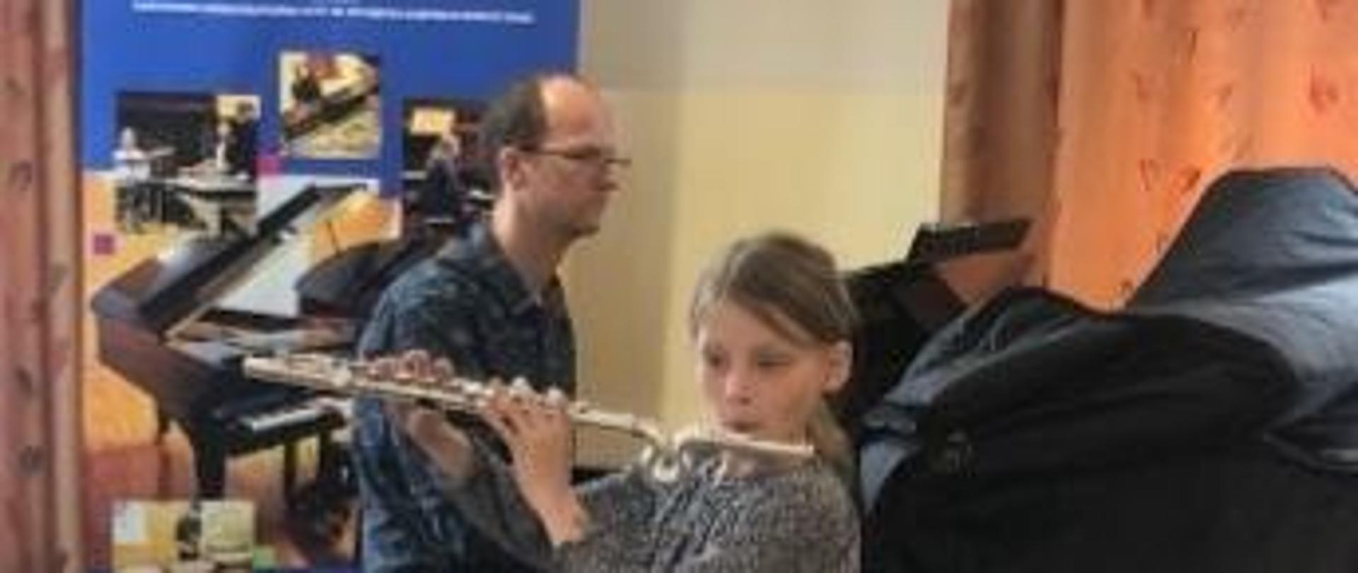 Na głównym planie dziewczynka grająca na flecie za nim pan akompaniator grający na czarnym fortepianie. W tle od lewej strony baner reklamowy unijny, okno z dużą pomarańczową zasłoną. 