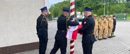 Na zdjęciu widoczny na placu zewnętrznym przed Komendą PSP w Gorlicach poczet flagowy przy maszcie w ubraniach reprezentacyjnych podczas podnoszenia flagi w zawiązku z uroczystością Dnia Flagi Rzeczypospolitej Polskiej. W tle zmiana służbowa w dwuszeregu w pozycji na baczność w ubraniach koszarowych koloru pisakowego i czarnego w czapkach.