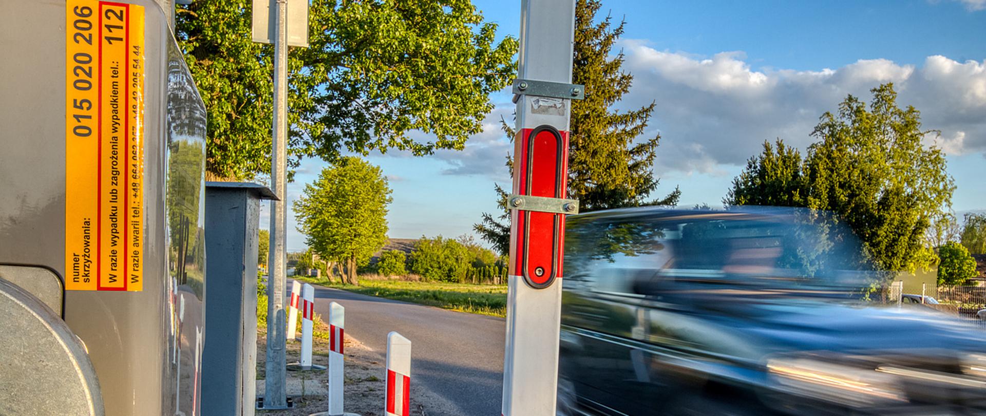 Na zdjęciu widać przejazd kolejowy i auto wjeżdżające na niego. W tle widać drogę i las. 
